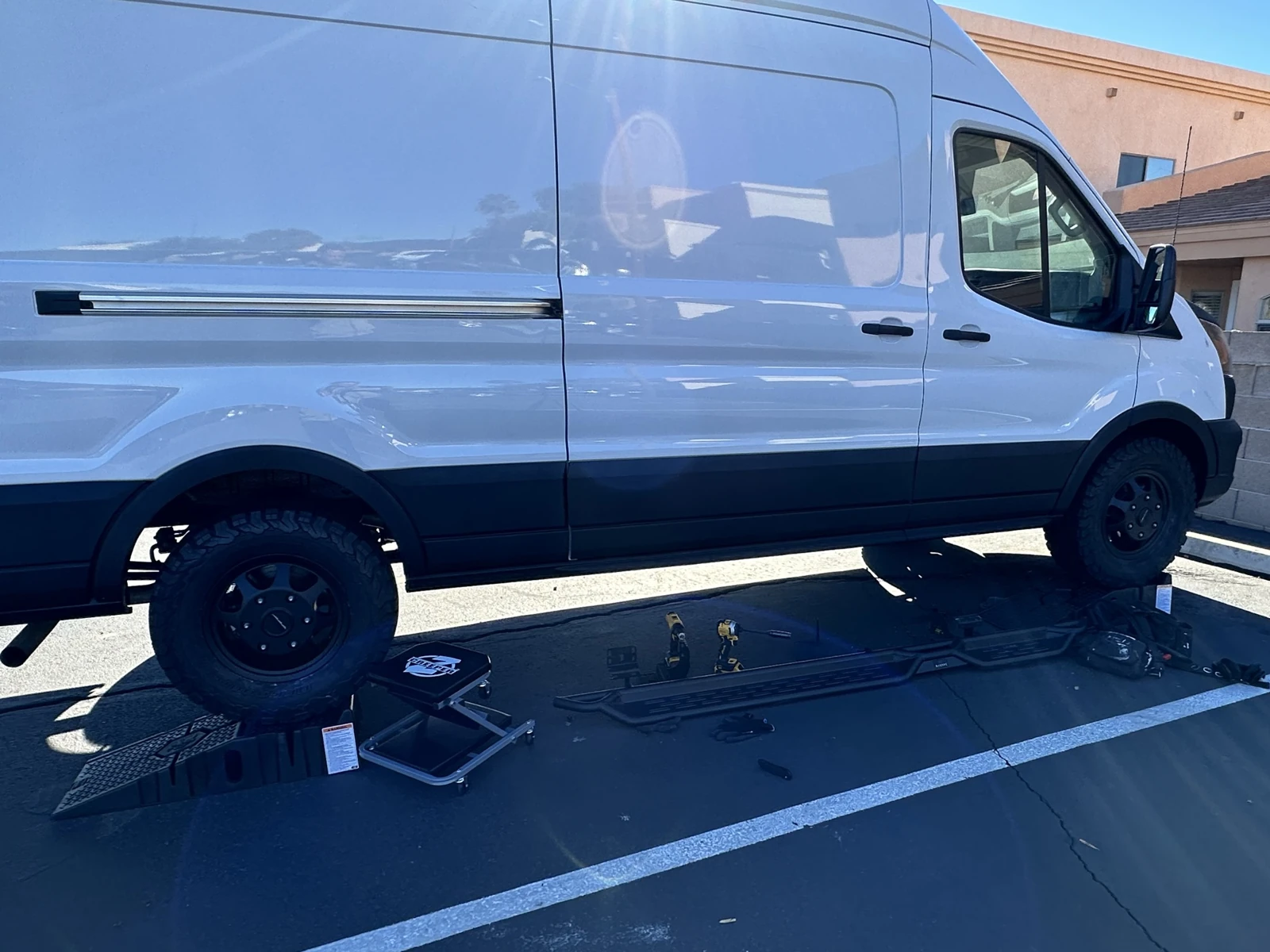 My Transit in the work area, with the passenger running board, mounting pieces, cordless tools and creeper seat laid out on the pavement.