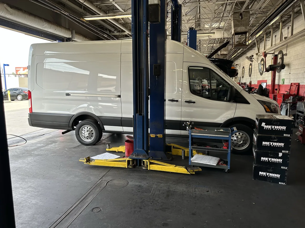 My white Ford Transit on its original silver wheels in a tire-shop bay beside Method wheel boxes.